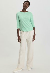 Haut long à manches vert clair, texturé et à encolure ronde, associé à un pantalon ample couleur crème et des baskets blanches.