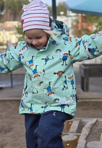 Ljusgrön regnjacka med färgglada serietidningsmönster, med figurer och djur. Mörkblå vattentäta byxor, leende barn som bär en randig hatt.
