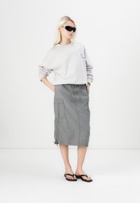 Felpa over-size grigia chiara con logo blu, abbinata a una gonna cargo grigia a lunghezza ginocchio con tasche e cordino. Infradito nere.