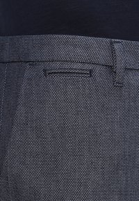 Mörkblått tyg med en subtil strukturerad mönster, med en slät front och en fickdetalj. Midjebandet är slätt och något elastiskt.