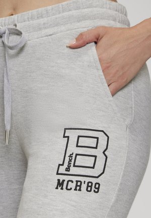 Hand in der Tasche einer hellgrauen Jogginghose mit Kordelzug und schwarzem "Bench B MCR '89"-Logo am Oberschenkel.