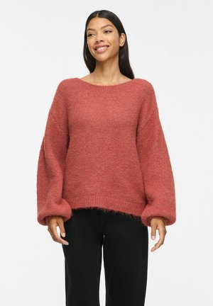 Femme souriante portant un pull ample texturé couleur rouille avec des manches ballon et un pantalon noir sur un fond blanc uni.