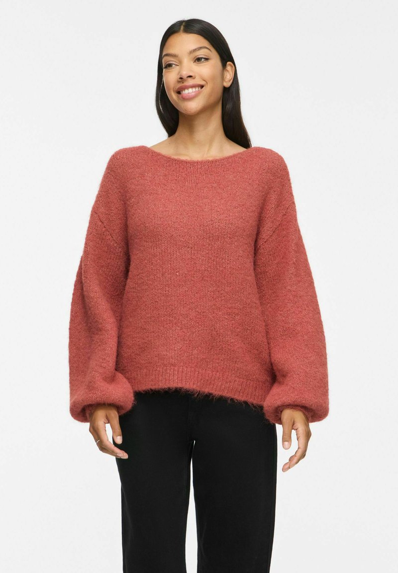 Femme souriante portant un pull ample texturé couleur rouille avec des manches ballon et un pantalon noir sur un fond blanc uni.