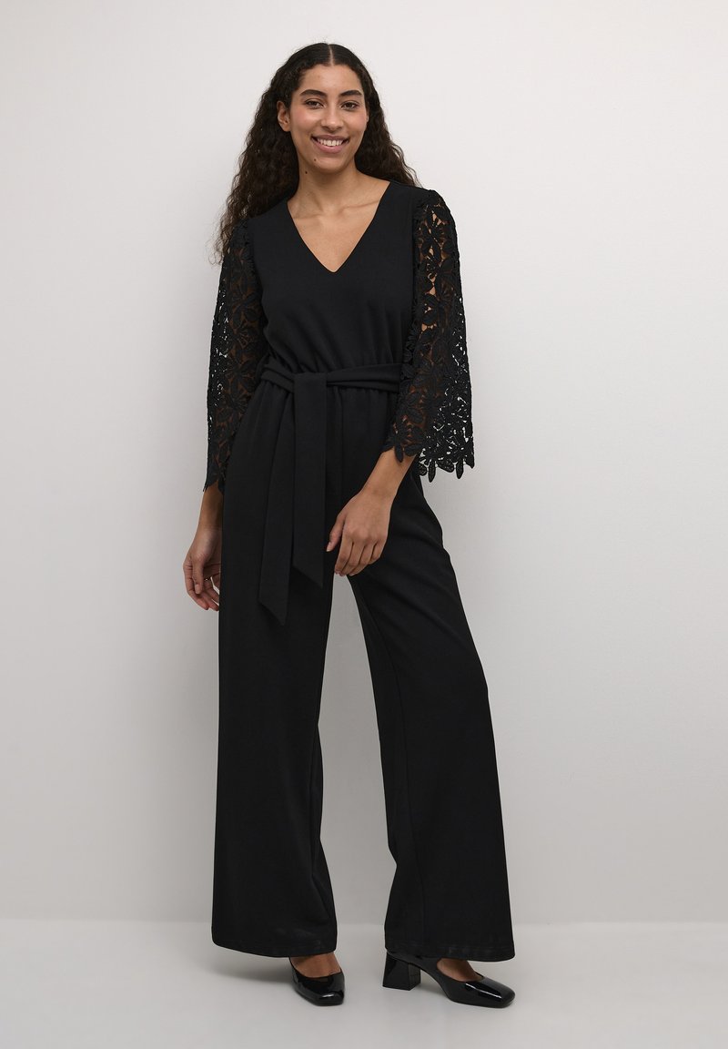 Completo nero con gambe ampie, scollatura a V e maniche a campana con pizzo. Presenta una vita con cintura e un tessuto morbido. Tacchi neri completano il look.