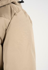 Giacca a vento beige con un tessuto strutturato e liscio. Presenta un logo in rilievo sulla manica e un cappuccio regolabile con chiusura a cordino.