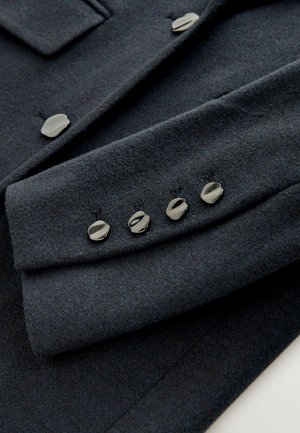 Donkergrijze wollen jas mouw met glanzende zilveren knopen en een gestructureerde afwerking. Bevat een zakflapdetail boven de knopen.