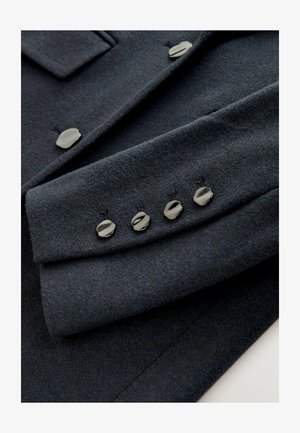 Dark gray wool jacket sleeve featuring smooth silver buttons and a textured finish. Includes a pocket flap detail above the buttons.