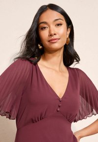 Bordová šaty s plisovanými priesvitnými rukávmi, výstrihom do V a tromi ozdobnými gombíkmi na poprsí. Hladká textúra, priliehajúci strih.