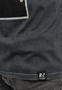 Grå T-shirt med en mjuk textur, med en svart grafisk tryck och en liten svart etikett vid fållen som står "R:̶C̶ RE:COVERED."