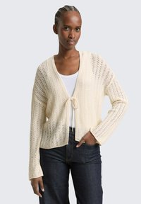 Vrouw met gevlochten haar, gekleed in een crèmekleurige gebreide cardigan die aan de voorkant is gestrikt, over een wit shirt en donkerblauwe spijkerbroek, met hand in de zak.