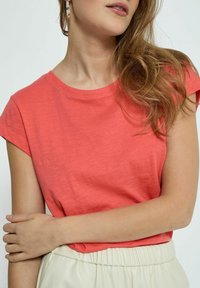Korálové tričko s krátkym rukávom a okrúhlym výstrihom, vyrobené z ľahkého materiálu. Modelka ho nosí so svetlo krémovými elastickými nohavicami.