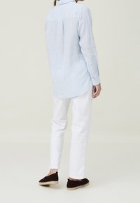 Camicia a righe azzurre chiare con maniche lunghe e polsini abbottonati. Pantaloni bianchi aderenti e espadrillas nere con suola intrecciata. Sfondo semplice.