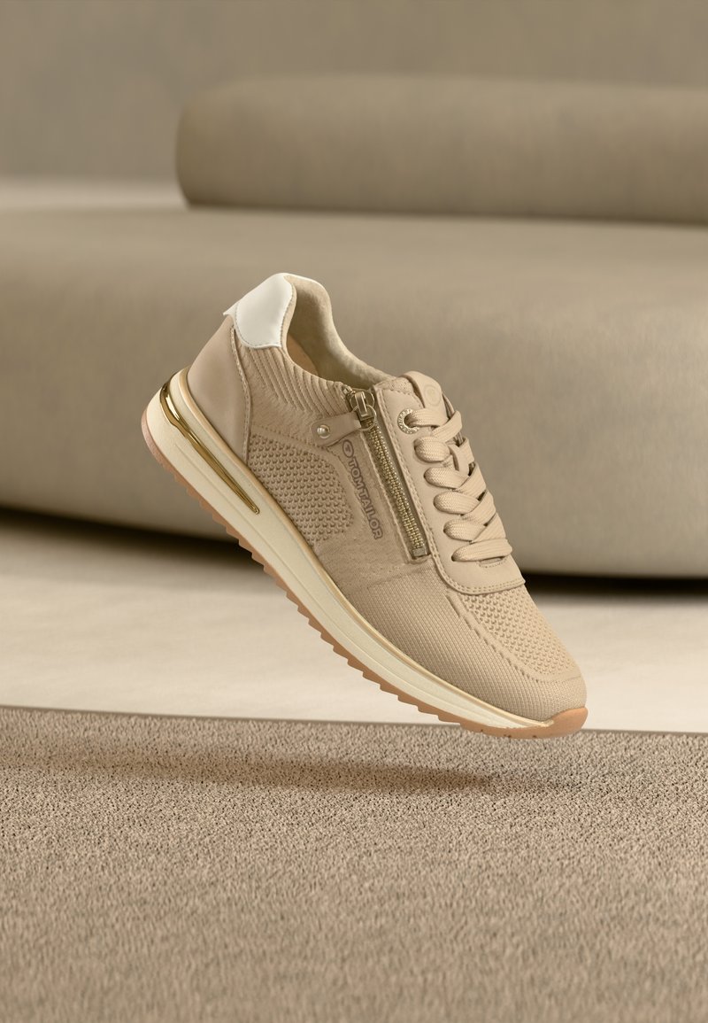 Baskets beige avec une tige texturée et respirante, un contrefort blanc, des accents dorés et une fermeture éclair latérale. Conception légère avec une semelle en caoutchouc.