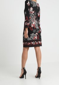 Kvinna klädd i en svart blommig knälång klänning med röda och vita mönster samt svarta högklackade sandaler med vristremmar.