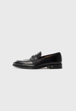 Black leather men's loafer with a decorative strap and low heel featuring a small metallic emblem on the side.