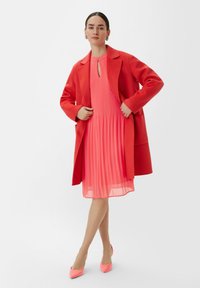 Czerwony płaszcz na plecioną różową sukienkę z okrągłym dekoltem i detalem w kształcie dziurki. Różowe czółenka ze spiczastym noskiem dopełniają stylizację.
