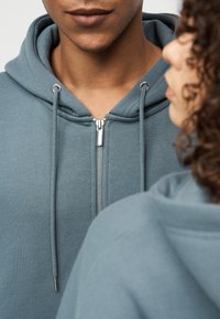 Φούτερ με κουκούλα και φερμουάρ σε τιρκουάζ χρώμα, με ασημένιο φερμουάρ και κορδόνια. Το ύφασμα φαίνεται μαλακό με υφή.