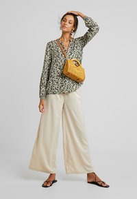 Blusa flor específica em verde oliva, calças largas creme e sandálias rasas pretas. A bolsa de palha amarela acrescenta textura e contraste de cor.