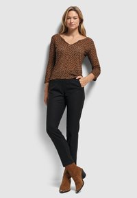 Kobieta stoi z jedną ręką w kieszeni, ubrana w brązową bluzkę w leopardzi wzór, czarne spodnie i brązowe zamszowe botki za kostkę na szarym tle.