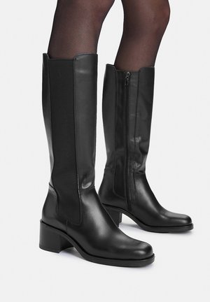 Czarne skórzane kozaki do kolan na słupku, z bocznym zamkiem błyskawicznym i elastyczną wstawką, noszone z przezroczystymi czarnymi rajstopami.