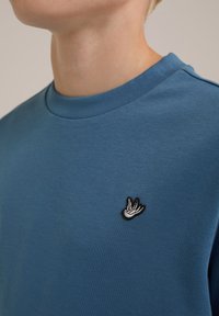 Blå bomullströja med rund halsringning och en liten broderad fågallogo på vänster bröst. Slät textur och minimalistisk design.