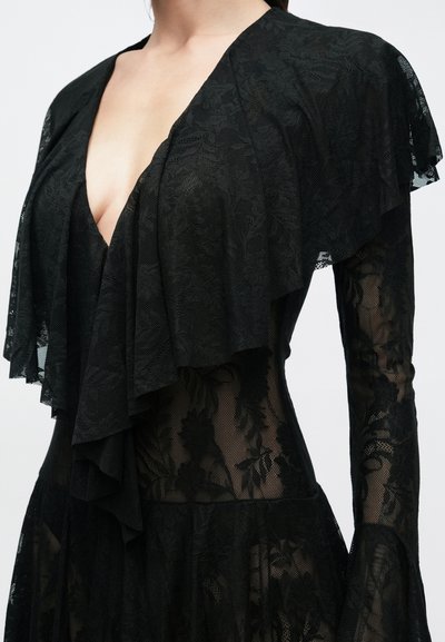 Γυναίκα που φοράει μαύρο διάφανο δαντελένιο φόρεμα με λουλουδάτα σχέδια και βαθύ V-λαιμό, με στρωμένες βολάν λεπτομέρειες στους ώμους και τα μανίκια.