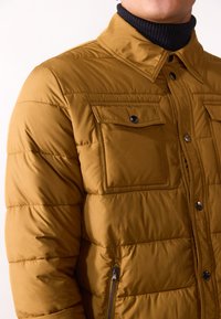 Brown padded jacket with a collar, featuring two chest pockets, a zippered pocket, and a textured black turtleneck underneath.