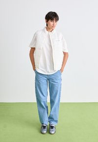 Persoon die staat met handen in de lichtblauwe spijkerbroekzakken, gekleed in een wit T-shirt met korte mouwen, een witte gewatteerde vest en zilveren sneakers op een groene vloer.