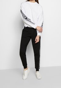 Bílá mikina se černým brandingem na rukávech, spárovaná s černými kalhotami se bočními pruhy a bílými teniskami.