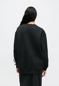 Μαύρο φούτερ με άνετη εφαρμογή, που διαθέτει λευκές ρίγες κατά μήκος των μανικιών και στρογγυλή λαιμόκοψη. Μαλακό, υφασμένο ύφασμα.