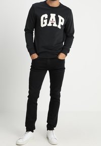 Svart bomullströja med vitt "GAP"-logotyp och röda accenter. Kombinerad med svarta jeans och vita sneakers, med enkelt mönster och passform.