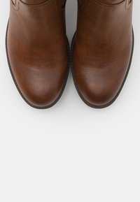 Bottes basses en cuir marron à texture lisse, bout arrondi, et détails de couture discrets. Aucun élément métallique ou motif visible.