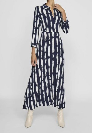 Kobieta ubrana w granatową sukienkę maxi z białymi pionowymi wzorami pędzla, z rękawami do połowy przedramienia i jasnobeżowymi botkami za kostkę.