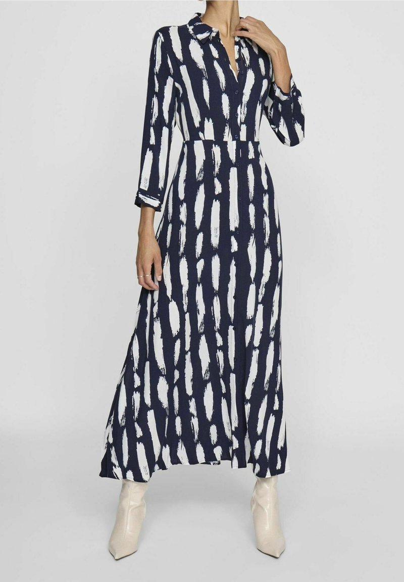 Femme portant une robe longue bleu marine avec des motifs de coups de pinceau blancs verticaux, des manches trois-quarts et des bottines beiges claires.
