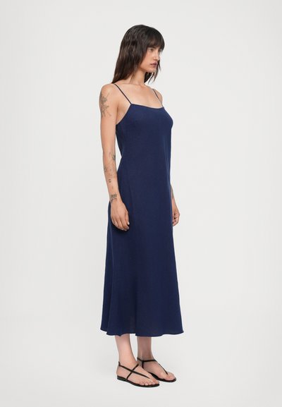 Vestido maxi sin mangas de color azul marino con tirantes delgados, tejido fluido y silueta recta. Un par de sandalias planas negras son visibles.