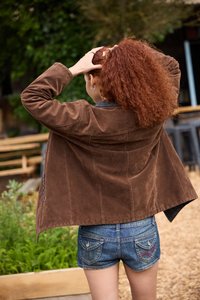 Braune Wildlederjacke mit lockerer Passform und schwarzen Lederelementen, kombiniert mit blauen Jeansshorts mit dekorativen Gesäßtaschen.