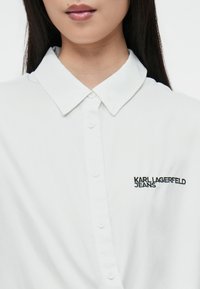 Camisa blanca de botones con cuello, con el logo bordado "KARL LAGERFELD JEANS" en negro. Textura de tela suave.