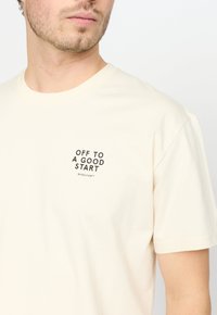 Crème katoenen T-shirt met een zwart gedrukt tekstgrafiek "OFF TO A GOOD START" op de linkerborst, met korte mouwen en een ronde hals.