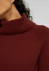 Pull en maille bordeaux avec un col côtelé haut et un détail texturé aux épaules, présentant une finition lisse et un design ajusté.