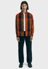 Jeune personne debout, portant une chemise à carreaux orange, marron et noir sur un t-shirt blanc, un pantalon noir et des chaussures noires.