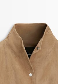 Límec béžové semišové bundy s našitými detaily a dvěma stříbrnými patentkami na světlém pozadí.