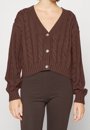 Femme portant un cardigan marron en tricot torsadé avec des boutons, à manches longues, et un pantalon sombre ajusté marron.