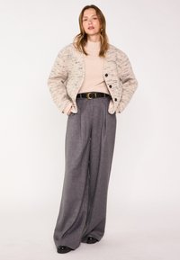 Veste texturée beige avec des éclats marins, boutons noirs ; pull beige clair ; pantalons gris à jambes larges ; ceinture noire avec boucle ronde ; chaussures noires.