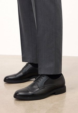 Zapatos de vestir de cuero negro con una textura suave, punteras redondeadas y un diseño de puntera cosida. Combinados con pantalones grises a medida.