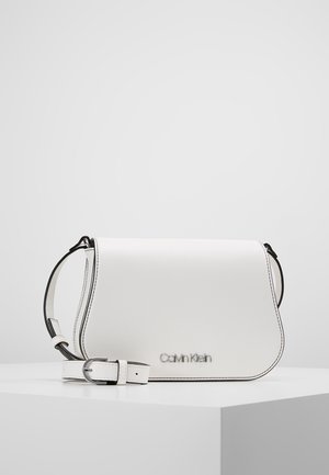 Sac bandoulière blanc Calvin Klein avec logo argenté, bords garnis de noir, et bandoulière réglable avec boucle sur une surface blanche avec un fond uni.