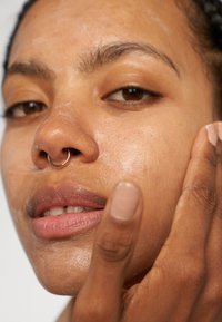 Close-up van het gezicht van een persoon met mediumbruine huid, een gouden septumring en glanzende huid, die wordt aangeraakt door een lichtbruine hand.