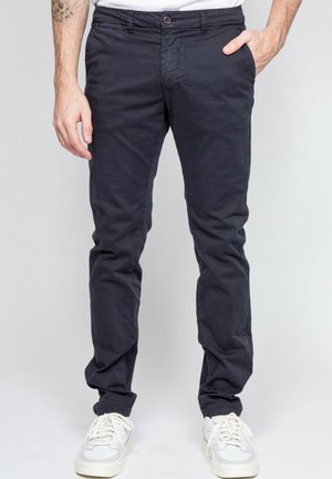 Homme portant un pantalon noir cintré avec des baskets blanches, une main dans la poche et un tatouage visible sur l'avant-bras gauche.
