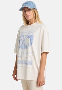 Kvinde iført en lys oversized T-shirt med et blåt palmetræprint og tekst, parret med cremefarvede bukser og en lyseblå denimkasket.