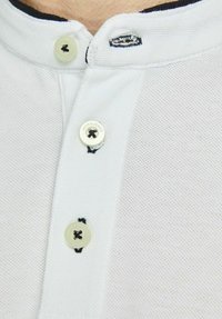 Witte polo met een textuur, ademend stof. Heeft een marineblauwe kraag, drie knopen en een klein logo op de linkerknoop.