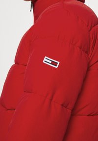 Giacca a vento rossa con texture imbottita, colletto alto e una patch con logo sulla manica. Chiusura con zip sul collo.